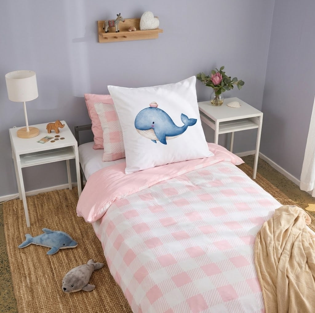 A children's single bed styled in a bedroom, featuring a pink and white checkered duvet cover, a matching pink pillow, and a white continental pillow with a blue whale illustration. The bed is flanked by white side tables with a lamp and a vase of flowers, set against a lavender wall.