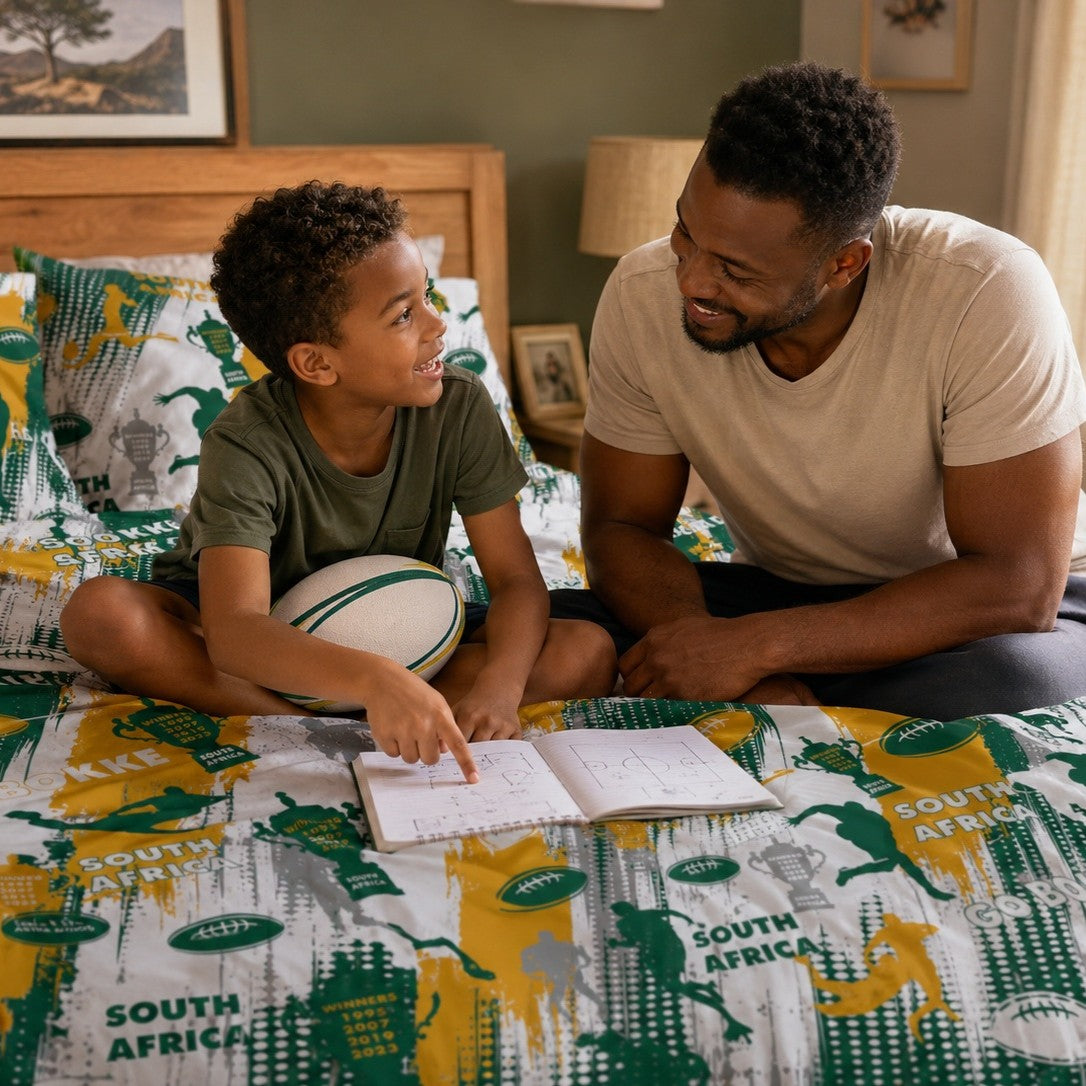 A father and son sitting together on a bed with a South Africa-rugby themed duvet, looking at a notebook. The boy holds a rugby ball, and the bed is decorated with matching pillows and a duvet cover featuring green and yellow sports graphics and text.