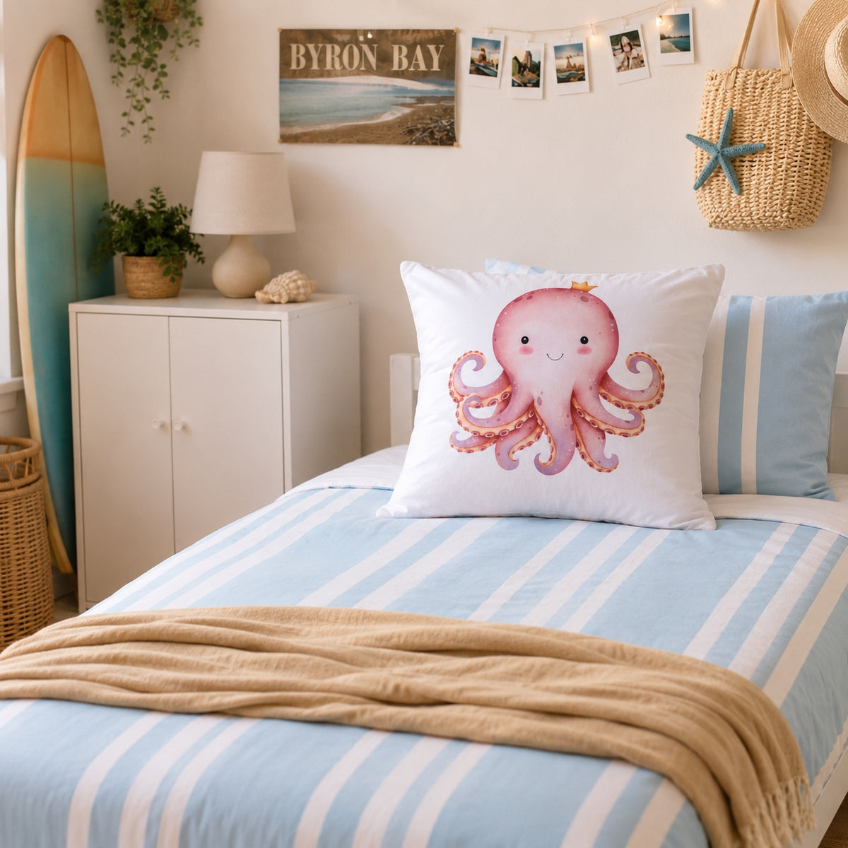 A styled bedroom scene with a bed made up in light blue and white stripes, a beige throw blanket, and a white continental pillow featuring a pink octopus illustration. A white cabinet, lamp, surfboard, woven bag, and wall decor are visible in the room.