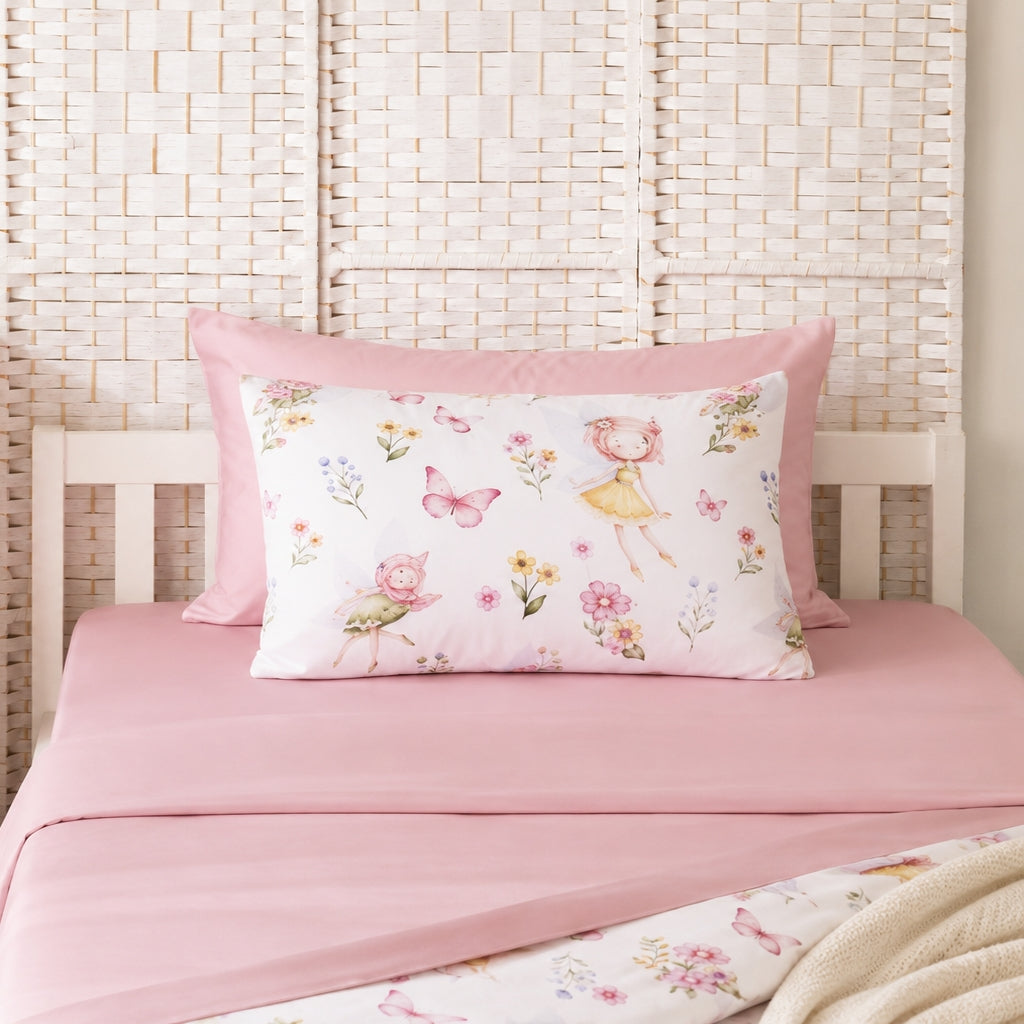 A pink bedding set on a bed, featuring a solid pink sheet and a white pillowcase printed with watercolor fairy and floral motifs, styled against a woven headboard.