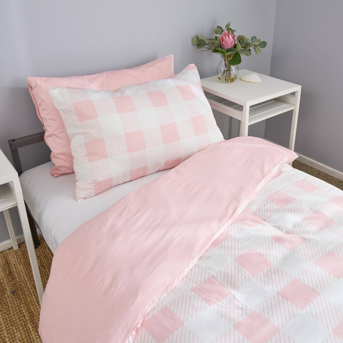 Pink and white checkered bedding set on a bed with a side table and flowers in the background.