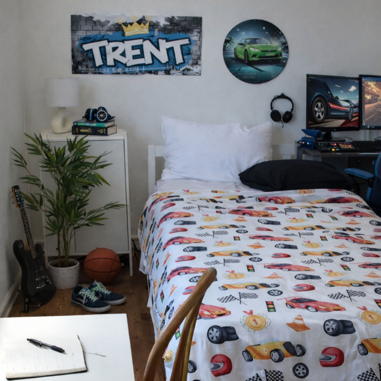 A bed in a bedroom featuring a white car-print bedding set with various sports cars. The room also contains a white nightstand with a lamp and camera, a potted plant, a basketball, two guitars, and a desk with computer monitors.