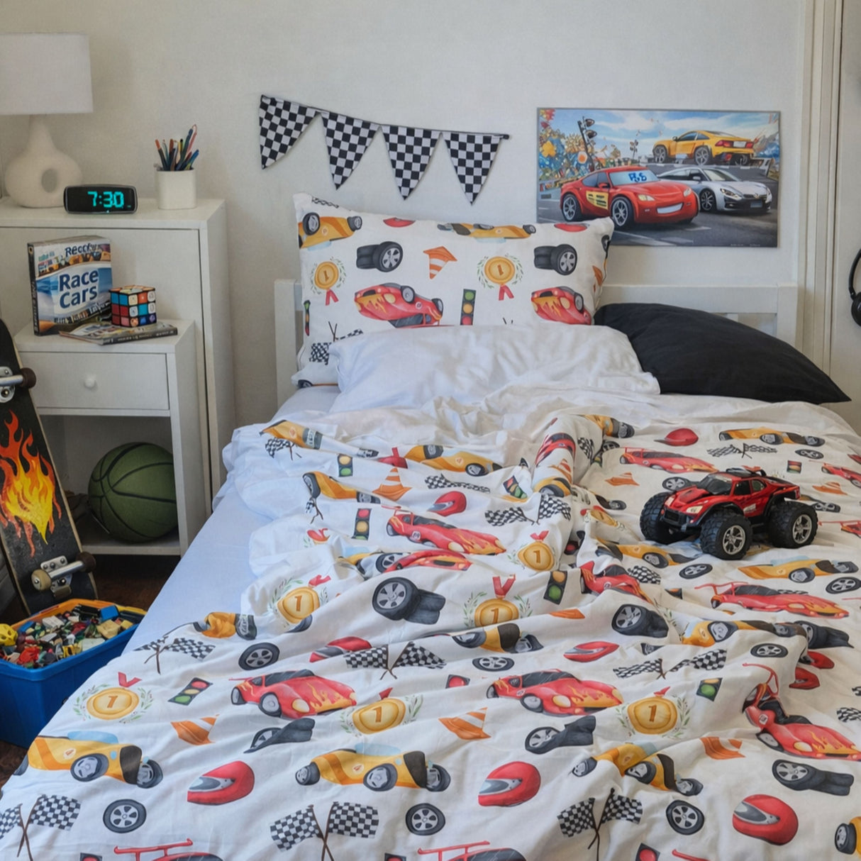 A children's bed in a bedroom styled with a white racing-themed bedding set featuring cartoon race cars, checkered flags, and racing motifs. A red monster truck toy sits on the bed, with a nightstand, lamp, and wall art visible in the room. Jo's Dreamland family designed organic kids bedding South Africa