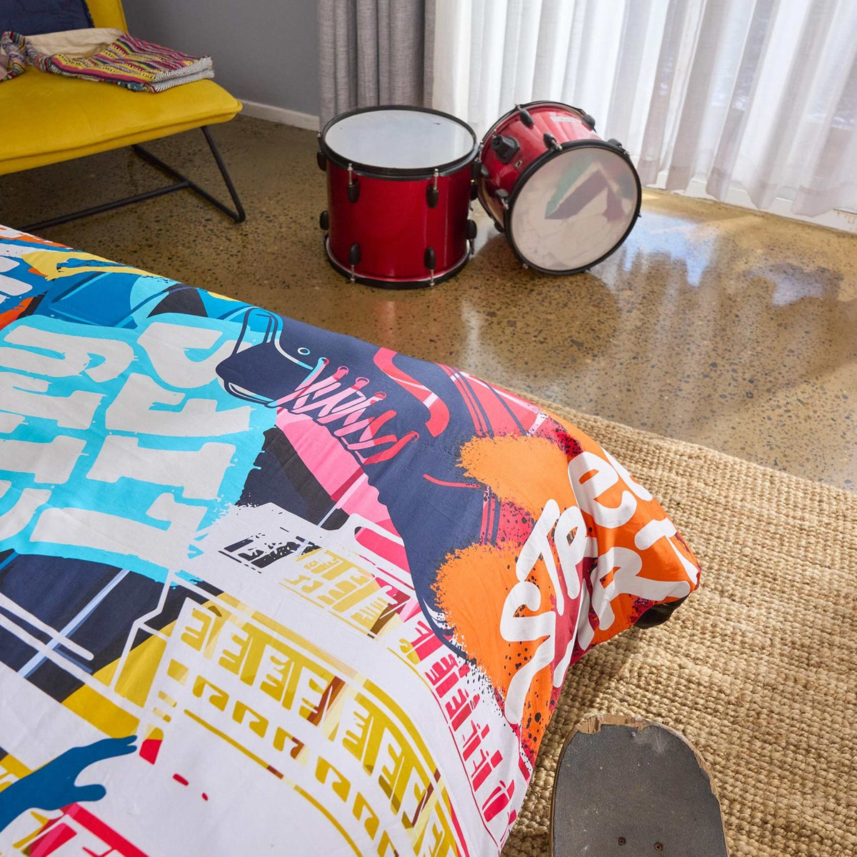 Colourful hip hop themed bedding close up on a bed, next to red drums and a yellow chair.