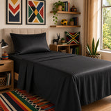 A neatly made bed dressed in black organic cotton bedding, including a black pillowcase, fitted sheet and top sheet, styled in a warm, modern bedroom with wooden shelves, framed art, plants, carpets.