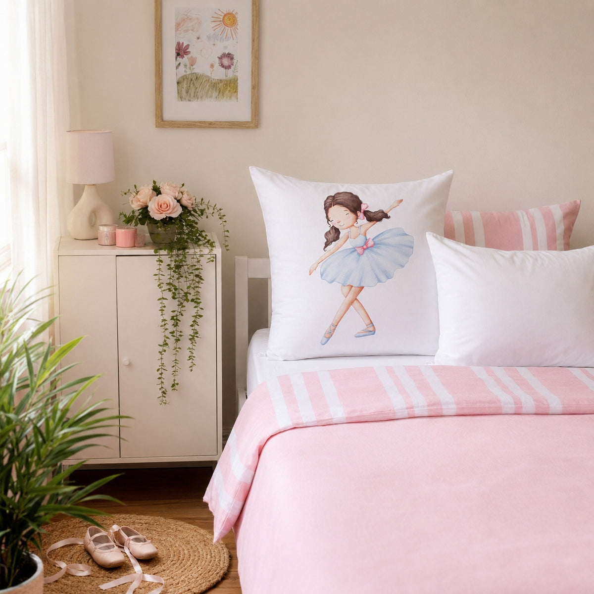 A white continental pillow featuring an illustration of a ballerina in a blue tutu, styled on a bed with pink and white striped bedding in a soft, sunlit room.