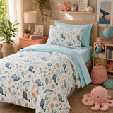 A children's bedding set styled on a bed in a decorated bedroom. The set includes a duvet cover and a matching pillowcase featuring a whimsical ocean-themed print with blue whales, sea urchins, starfish, and coral on a white background, accented by solid light blue sheets and pillowcases.