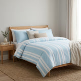 A light blue and white striped bedding set with matching pillows and a beige accent pillow, styled on a wooden bed frame in a bright, modern bedroom.
