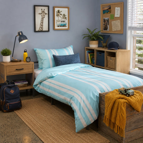 A styled bedroom scene showing a bed dressed in a light blue and white striped duvet cover with matching pillows, alongside a navy blue accent pillow. The room features a light blue wall, wooden shelving, a bedside table with a lamp, and a window with blinds.