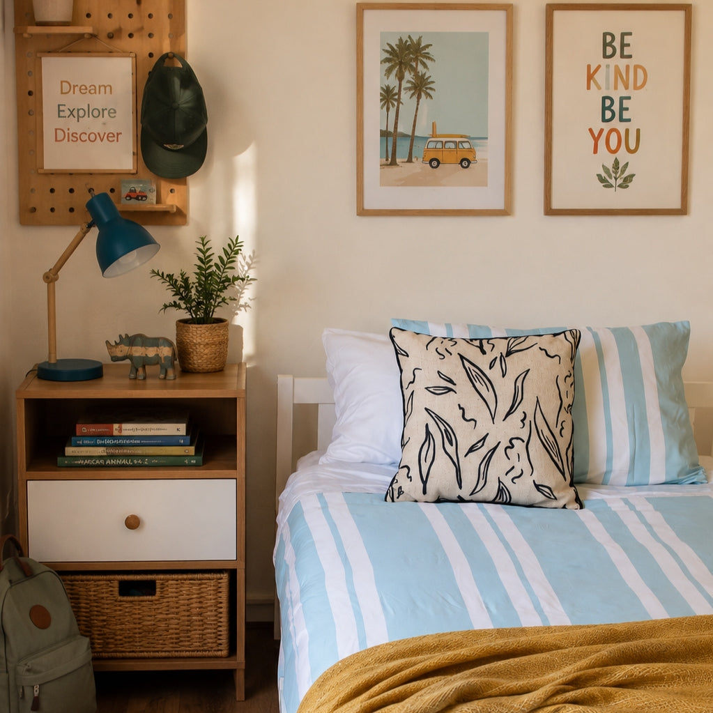 A styled bedroom corner with a bed dressed in light blue and white striped bedding, a patterned accent pillow, a wooden nightstand with books and a blue lamp, wall art, and a pegboard with a cap.