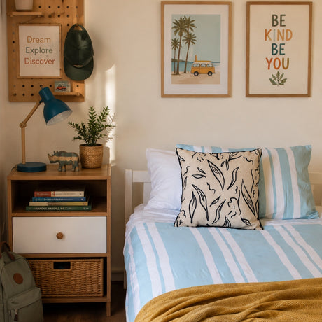 A styled bedroom corner with a bed dressed in light blue and white striped bedding, a patterned accent pillow, a wooden nightstand with books and a blue lamp, wall art, and a pegboard with a cap.