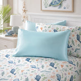 A neatly made bed in a bright bedroom featuring a light blue pillowcase in front of a white duvet cover printed with blue whales, coral, and sea plants. Matching patterned pillows rest behind it against a white headboard, with a bedside lamp, plant, and framed turtle artwork visible.