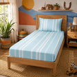 A light blue and white striped fitted sheet set on a wooden single bed frame, styled in a warm, decorated bedroom with plants, a shelf, and wall art.