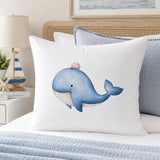 A white continental pillow featuring a watercolor illustration of a blue whale wearing a small striped hat, styled on a bed with blue bedding and decorative pillows.
