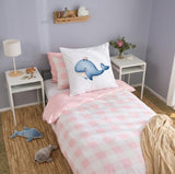 A children's single bed styled in a bedroom, featuring a pink and white checkered duvet cover, a matching pink pillow, and a white continental pillow with a blue whale illustration. The bed is flanked by white side tables with a lamp and a vase of flowers, set against a lavender wall.