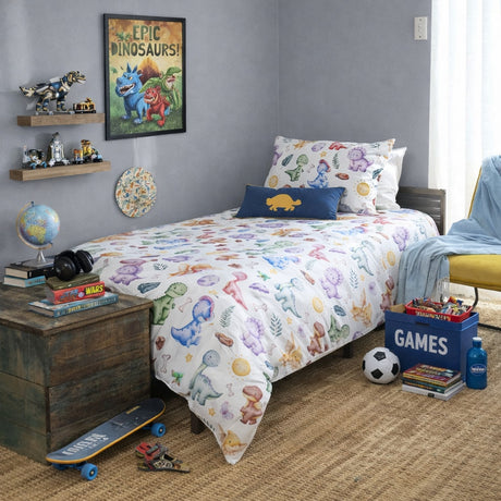 A kids' bedroom scene featuring a bed dressed in a white comforter and pillow shams printed with colorful dinosaur illustrations. The room includes a gray wall, wooden shelves with toys, a framed 'Epic Dinosaurs' poster, a nightstand with books and a globe, and a yellow chair with a blue throw blanket.