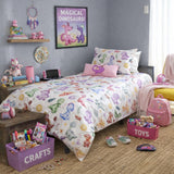 A children's bedroom scene with a bed dressed in a colorful dinosaur-print kids duvet cover and matching pillowcase. The room includes a pink backpack, purple toy bin, shelf with plush toys, wall art reading 'Magical Dinosaurs!', and a gray wall background.