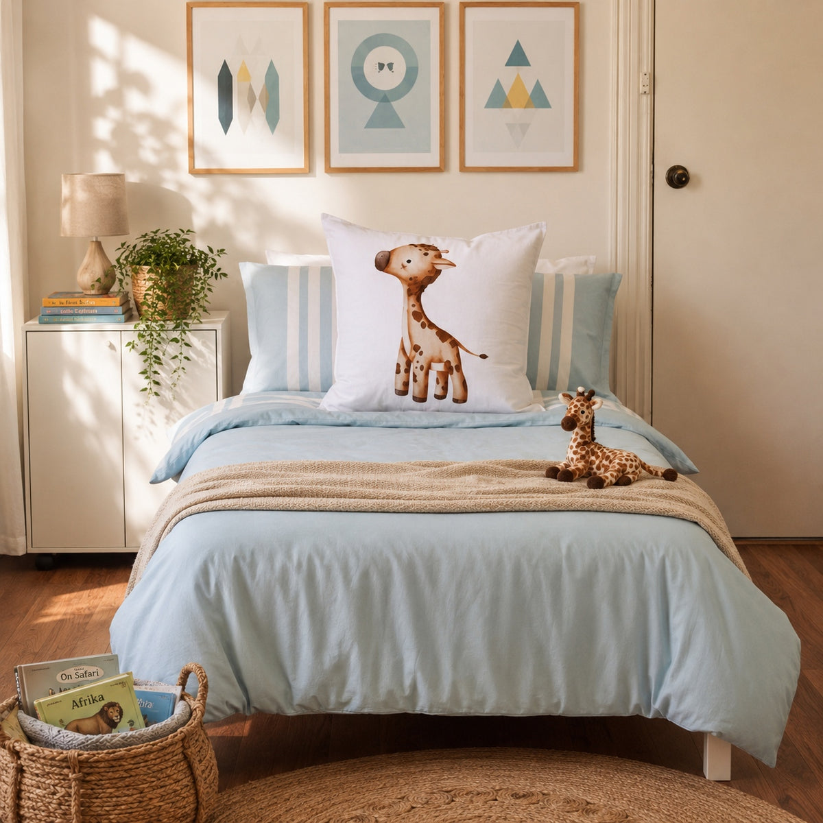 A styled children's bedroom featuring a light blue bedding set on a single bed, including a duvet cover, matching pillow shams, and a central white continental pillow with a printed giraffe. The room is decorated with three framed geometric wall art prints, a small white side cabinet with a lamp and books, and a wicker basket on the floor.