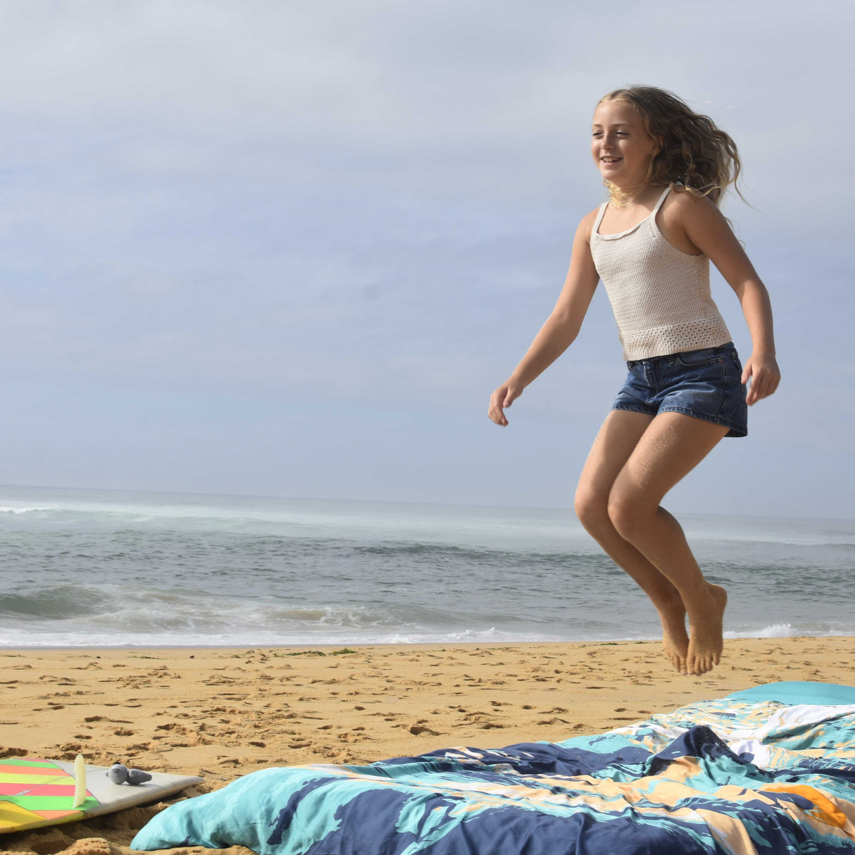 A young girl smiling and jumping on a sandy beach, wearing a cream knit tank top and denim shorts. A colorful beach blanket is spread on the sand in the foreground, with the ocean and cloudy sky behind her.