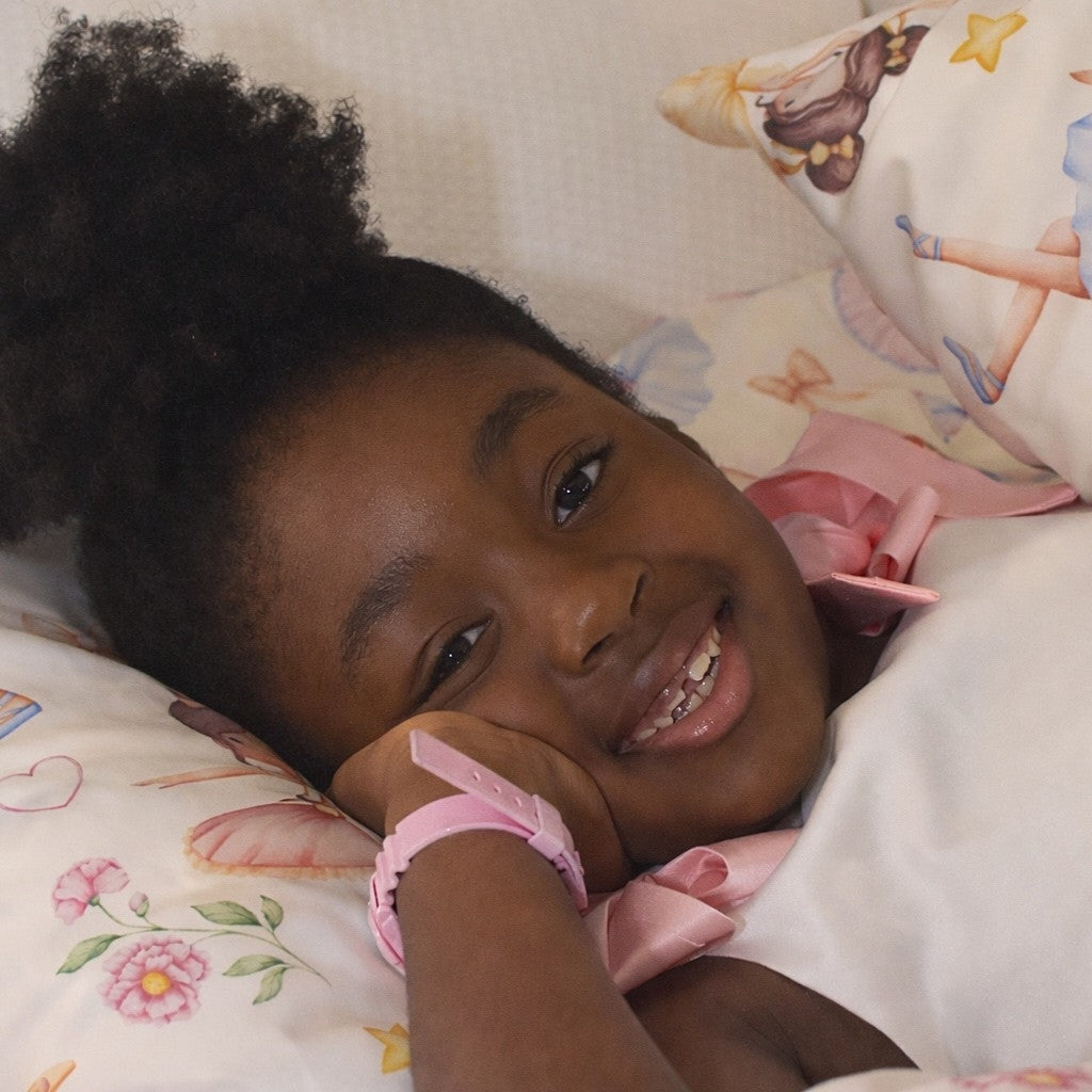 A young child with dark curly hair smiling while lying in bed, wearing a pink watch and resting on a white pillow with a colourful  ballerina print.