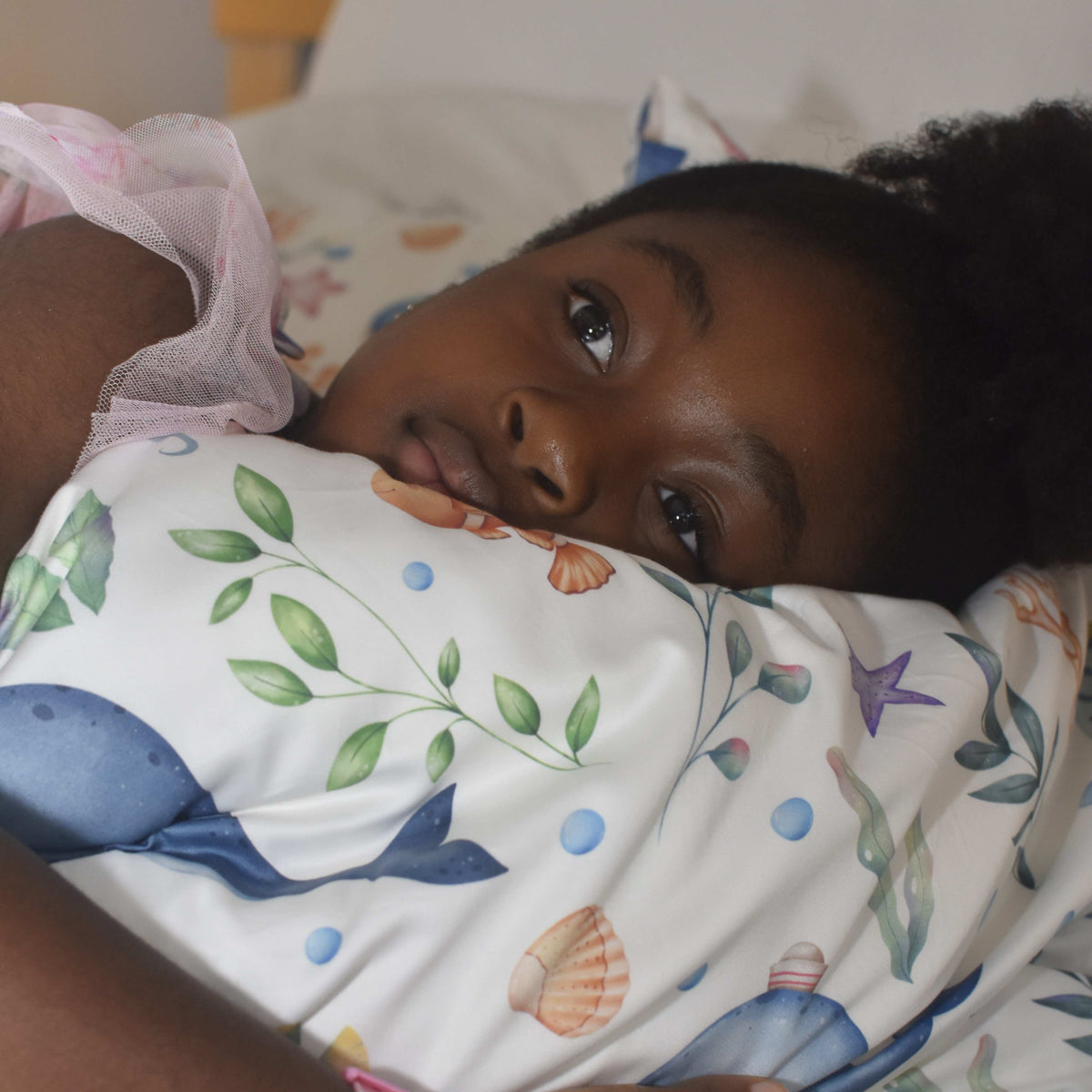 A young child lying on a bed, resting their head on a white pillowcase printed with a colorful undersea theme including whales, seashells, and green leaves.