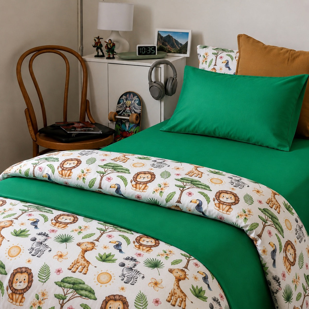 A styled bed in a kids bedroom setting featuring a green solid sheet and pillowcase layered with a white kids duvet cover printed with safari animals like lions, giraffes, zebras, and toucans. The room includes a wooden chair, a white side table with a lamp and headphones, and a mustard yellow pillow.