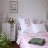 A styled bedroom scene showing a bed with pink and white striped bedding, a white pillow featuring a watercolor illustration of a fairy, a folded green blanket, a white nightstand with a lamp and flowers, and a framed floral print on the wall.