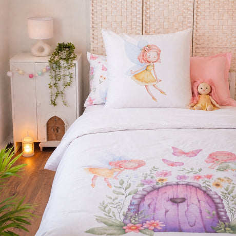 A styled bed in a nursery setting featuring a white fairy-themed bedding set with a purple fairy door, pink butterflies, and a white continental pillow with a pink-haired fairy illustration. The room includes a white nightstand with a lamp and trailing plant, a woven headboard, and a small doll.