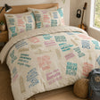 A bed dressed in a printed motivational duvet cover and matching pillowcases featuring motivational quotes in pink, blue, and gray over a cream background, styled in a bedroom with a wooden headboard, bedside lamp, guitar, and a backpack on the floor.