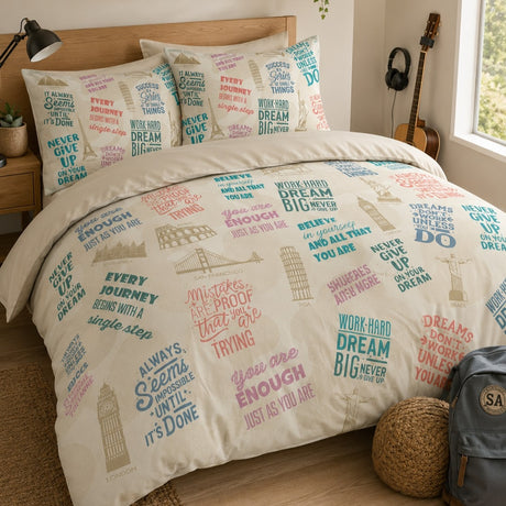 A bed dressed in a printed motivational duvet cover and matching pillowcases featuring motivational quotes in pink, blue, and gray over a cream background, styled in a bedroom with a wooden headboard, bedside lamp, guitar, and a backpack on the floor.