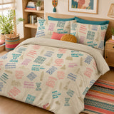 A bed dressed in a beige duvet cover and matching pillowcases featuring a colorful collage print of motivational phrases like 'NEVER GIVE UP', 'BELIEVE', and 'ALL THAT YOU ARE', styled in a warm, decorated bedroom with plants, a wooden headboard, and a patterned rug.