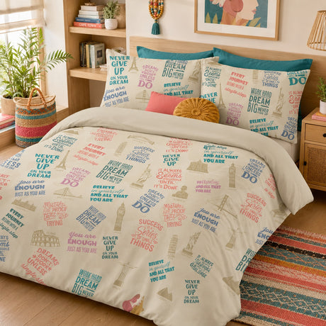 A bed dressed in a beige duvet cover and matching pillowcases featuring a colorful collage print of motivational phrases like 'NEVER GIVE UP', 'BELIEVE', and 'ALL THAT YOU ARE', styled in a warm, decorated bedroom with plants, a wooden headboard, and a patterned rug.