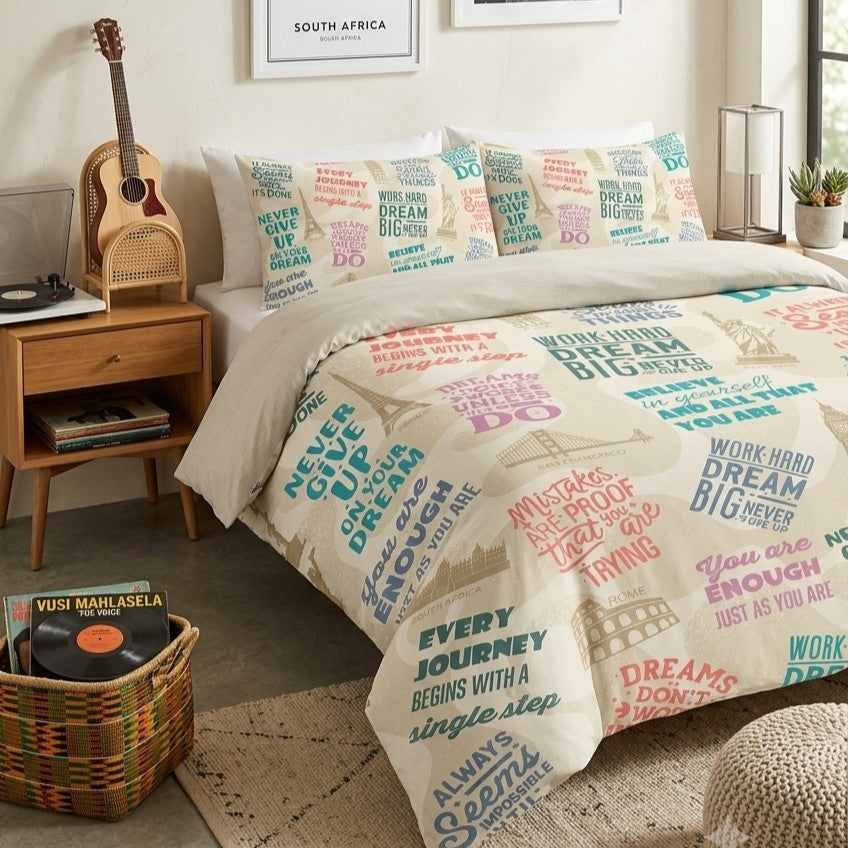 A bed dressed in a cream bedding set with colorful travel-themed typography and motivational quotes and illustrated motifs, styled in a bedroom with a wooden nightstand, guitar, basket, and framed wall art.