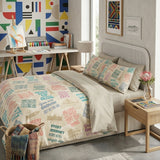 A beige bedding set with a colorful typographic motivational print is styled on a bed in a modern, brightly lit bedroom. The set includes a duvet cover and two matching pillowcases.