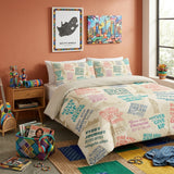 A bed dressed in a cream bedding set printed with colorful motivational text and travel-themed graphics, styled in a bedroom with peach walls, framed wall art, a wooden nightstand, and a window.