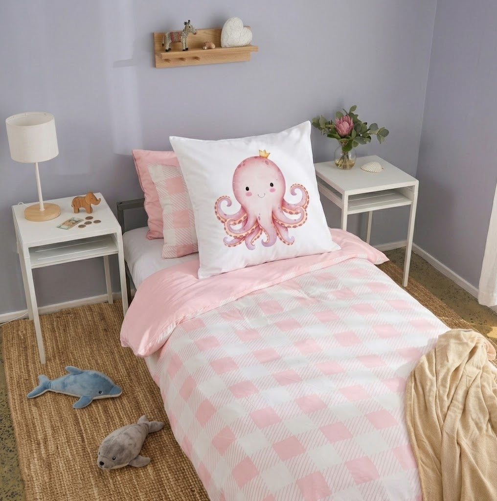 A styled children's bedroom featuring a single bed with a pink and white checkered duvet, a pink octopus continental pillow on a white pillow, and matching pink bedding. The room has lavender walls, white side tables with lamps and a vase of flowers, and a woven jute rug with plush toys.