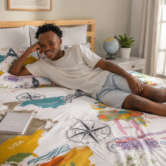 A teenager lies on a bed wearing a white t-shirt and light blue shorts, showcasing a colorful travel-themed duvet cover with world map graphics, compass roses, and travel stamps. The bedroom setting includes a wooden headboard, a white nightstand with a globe and plant, and a window with sheer curtains.