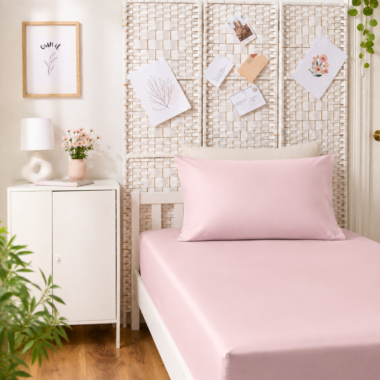 A light pink bedding set, including a fitted sheet and a matching pillowcase, styled on a bed in a bright, modern bedroom with a white nightstand, rattan headboard, and framed wall art.