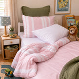 A children's bed in a bright bedroom, featuring a pink and white striped duvet cover, matching pillow shams, a white pillow, and an olive green back pillow. The bed is styled with a wooden nightstand, a star-patterned lamp, a small potted plant, and a teddy bear.