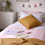 A cozy bedroom scene showing a bed styled with pink and white striped bedding, a mustard yellow textured pillow, white pillows, a white nightstand, and a small plant.