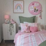 A cozy children's bedroom featuring a bed styled with pink and white striped bedding, a plush unicorn toy, and a pink heart-shaped pillow. The room includes a white side cabinet with a pink lamp, a framed rainbow print on the wall, and a pink circular bulletin board.