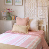 A neatly made bed in a softly styled bedroom, featuring a pink and white striped duvet cover, blush pink pillows, a cream pillow, and a beige fringed accent pillow. The background includes a woven beige headboard, a white sideboard with a vase of flowers and framed prints.