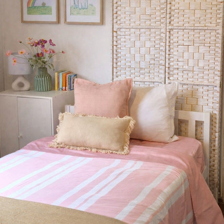 A neatly made bed in a softly styled bedroom, featuring a pink and white striped duvet cover, blush pink pillows, a cream pillow, and a beige fringed accent pillow. The background includes a woven beige headboard, a white sideboard with a vase of flowers and framed prints.