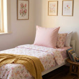 A styled bed in a softly lit bedroom featuring a white bedding set with a delicate pink and yellow floral and fairies print, a solid light pink continental pillow, a matching floral accent pillow, and a mustard yellow throw blanket.