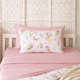 A pink bedding set on a bed, featuring a solid pink sheet and a white pillowcase printed with watercolor fairy and floral motifs, styled against a woven headboard.