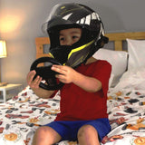 A child wearing a black and white full-face motorcycle helmet with yellow accents sits on a bed with a race car themed duvet cover , holding a black steering wheel prop.