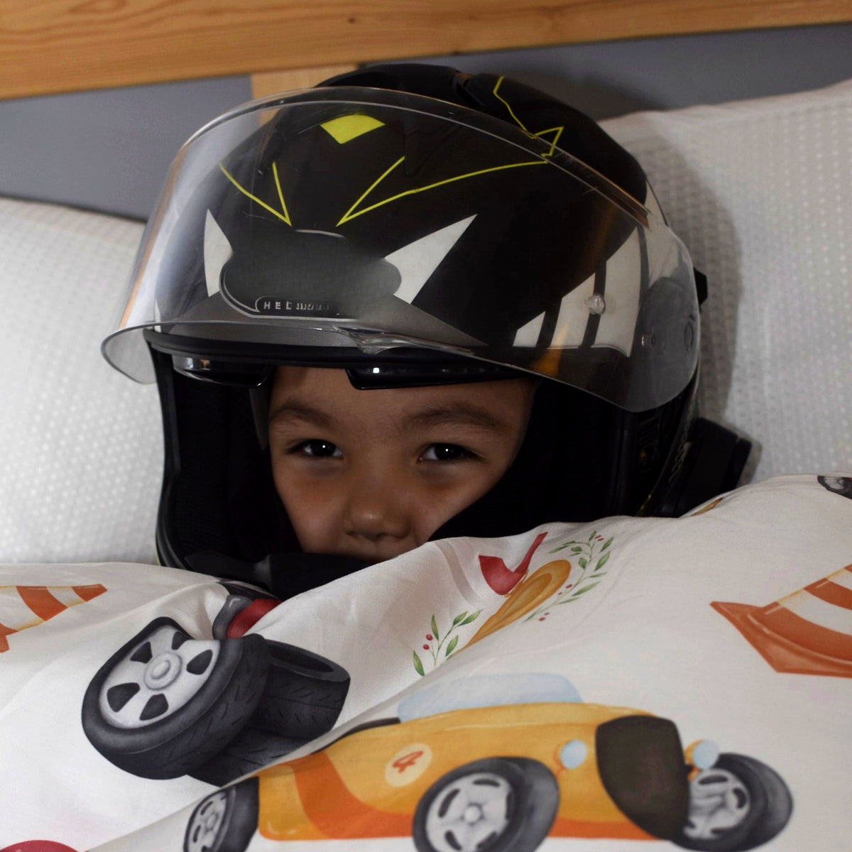A child wearing a black full-face motorcycle helmet with a clear visor, peeking out from under a white race car themed duvet cover with colourful race cars.