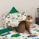 A brown tabby cat sits on a bed with a white duvet and matching pillowcase printed with colorful South African-themed graphics, including the word 'SAWKS' and green accents. A green pillow is visible behind the bed against a plain wall.