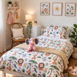 A children's bed styled in a bright room, featuring a safari-themed duvet cover and pillowcase with colourful animal prints including lions, giraffes, elephants, and birds. A pink plush elephant toy sits on the bed, with a wooden chair, bedside table, and framed wall art in the background.