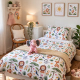 A children's bed styled in a bright room, featuring a safari-themed duvet cover and pillowcase with colourful animal prints including lions, giraffes, elephants, and birds. A pink plush elephant toy sits on the bed, with a wooden chair, bedside table, and framed wall art in the background.